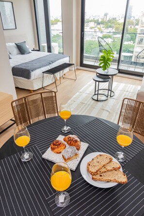 Habitació d'hotel moderna amb un llit gran, zona de dinar amb esmorzar i vista a la ciutat a través de grans finestres.
