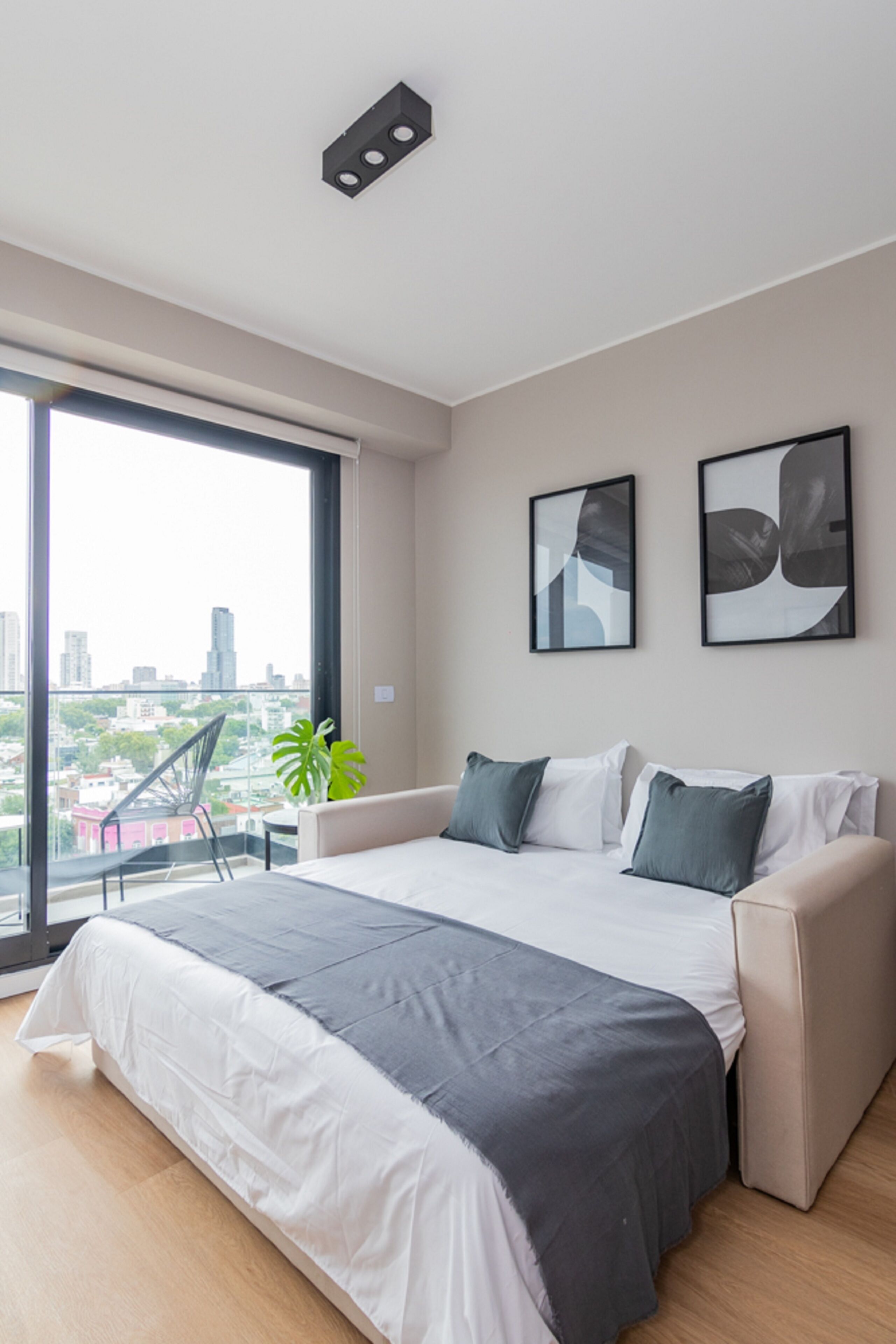 Une chambre moderne avec un grand lit, une vue sur la ville et un balcon agrémenté d’une chaise et d’une plante.
