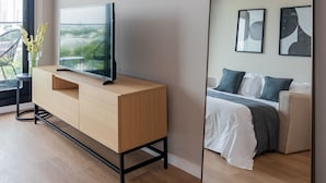 A modern hotel room with a large mirror, a flat-screen TV on a wooden cabinet, a bed with a gray and white duvet, and a window with a view of greenery. - Milenia Soho by iPPA (Buenos Aires)