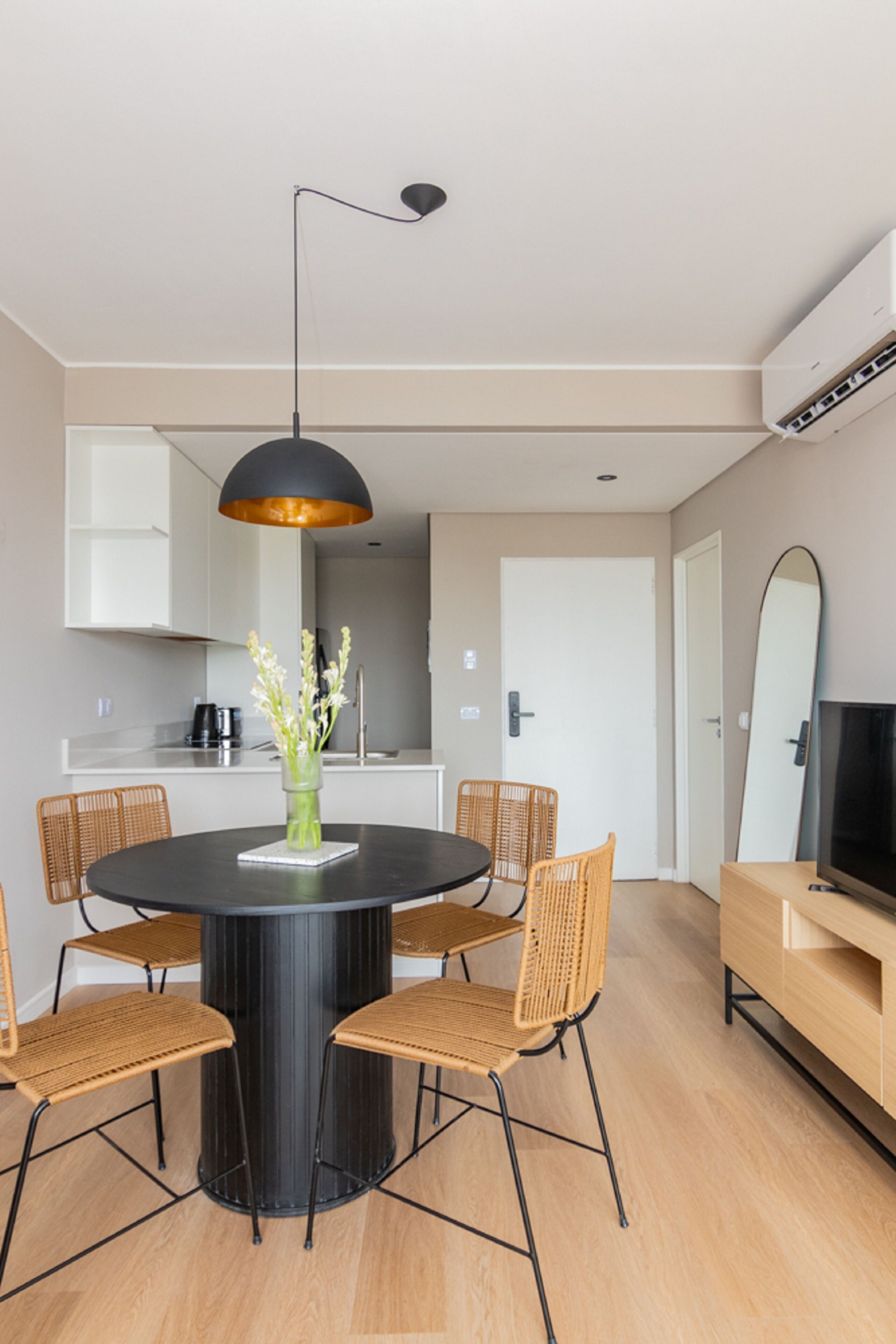 A modern dining area with a round table, six wicker chairs, a black pendant light, and a vase with flowers.
