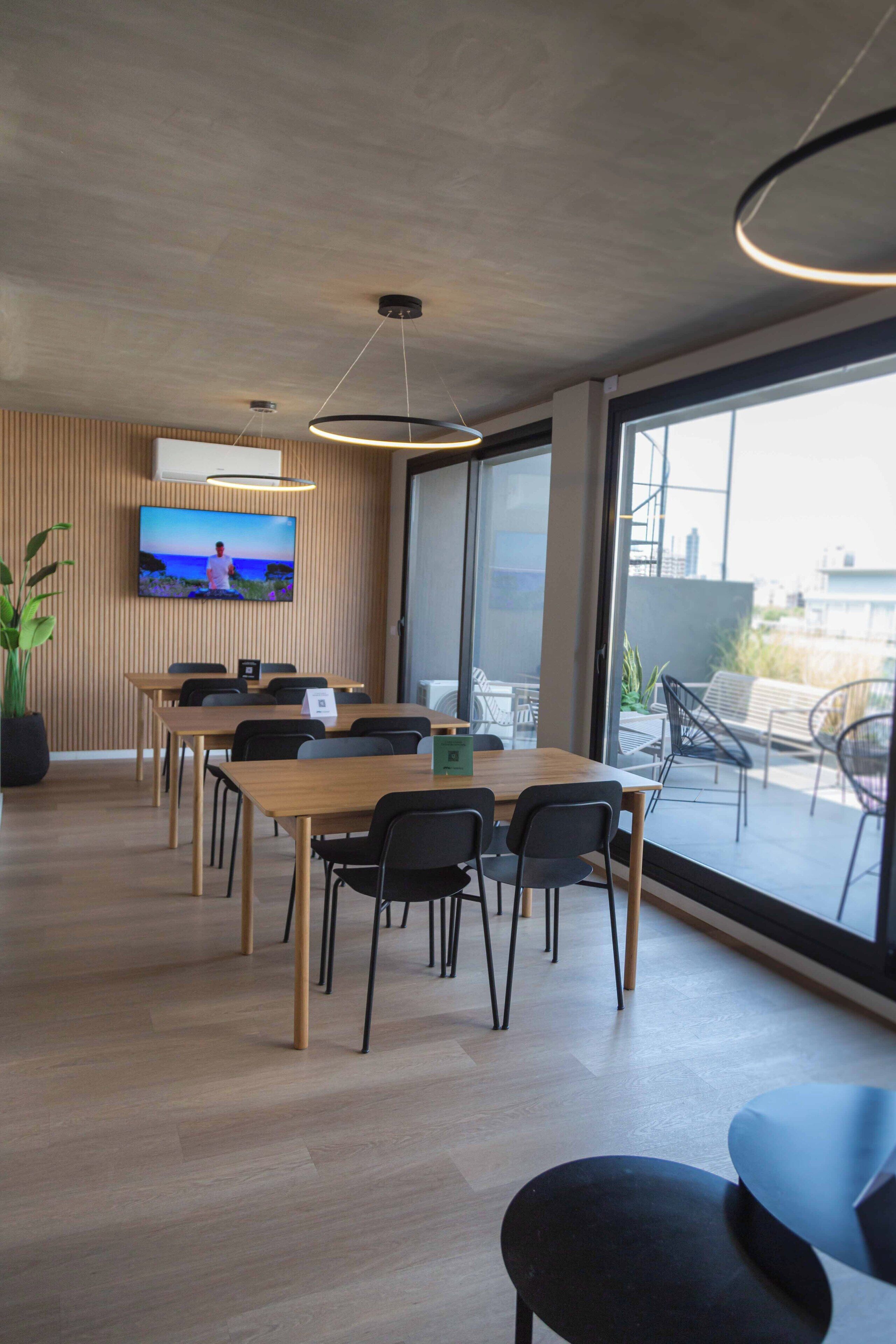 A modern conference room with a large table, chairs, a TV on the wall, and a view through large windows.