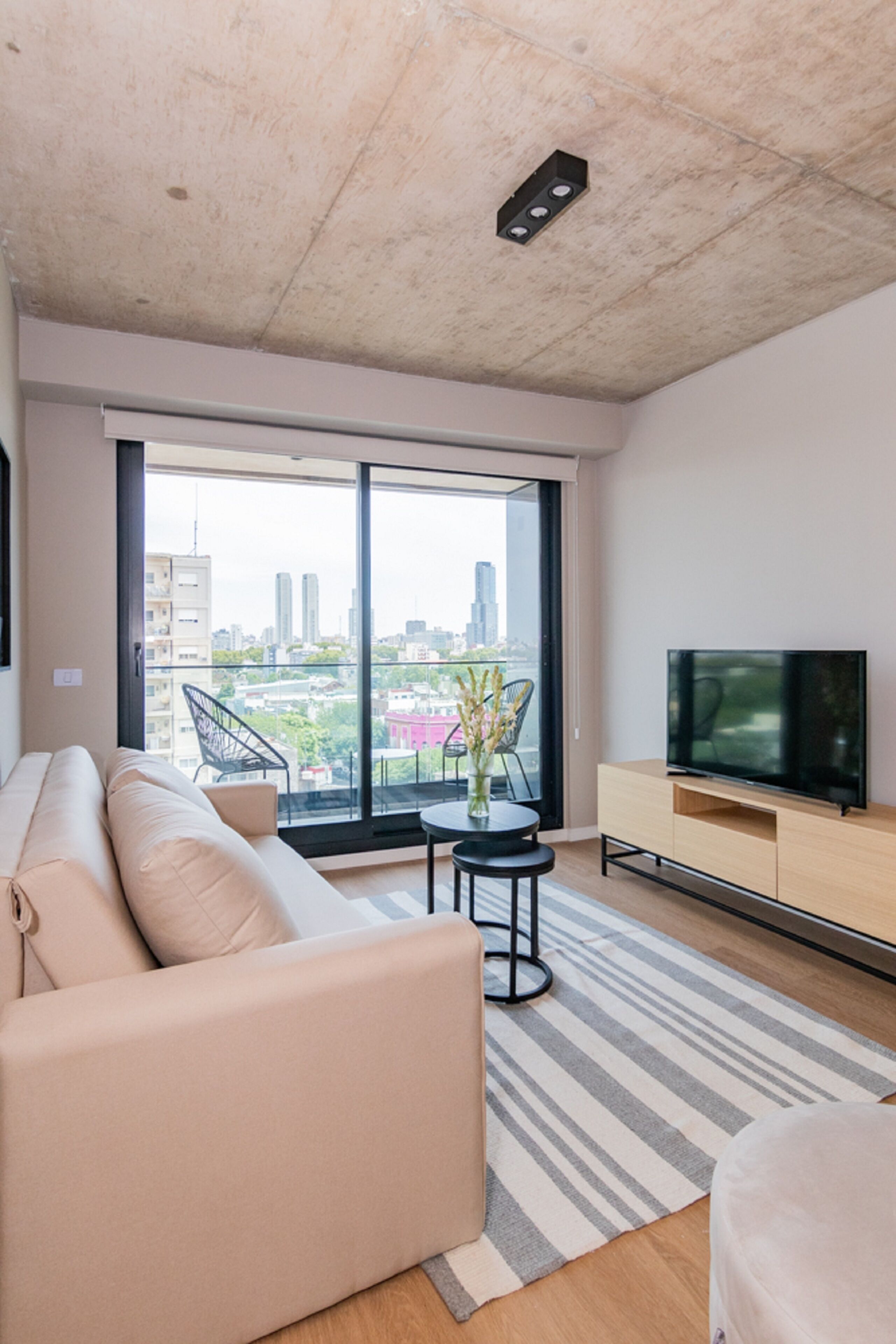 Una sala de estar moderna con un sofá, un televisor sobre un mueble de madera y un ventanal con vista a la ciudad.