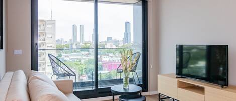 Una sala de estar moderna con un sofá, un televisor sobre un mueble de madera y un ventanal con vista a la ciudad.