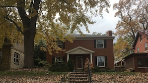 A two-story brick house with a front porch, a large tree in front, and a staircase leading up to the entrance. - Spacious 6BR, 10 bed Derby House. Walk to the track. (Louisville)