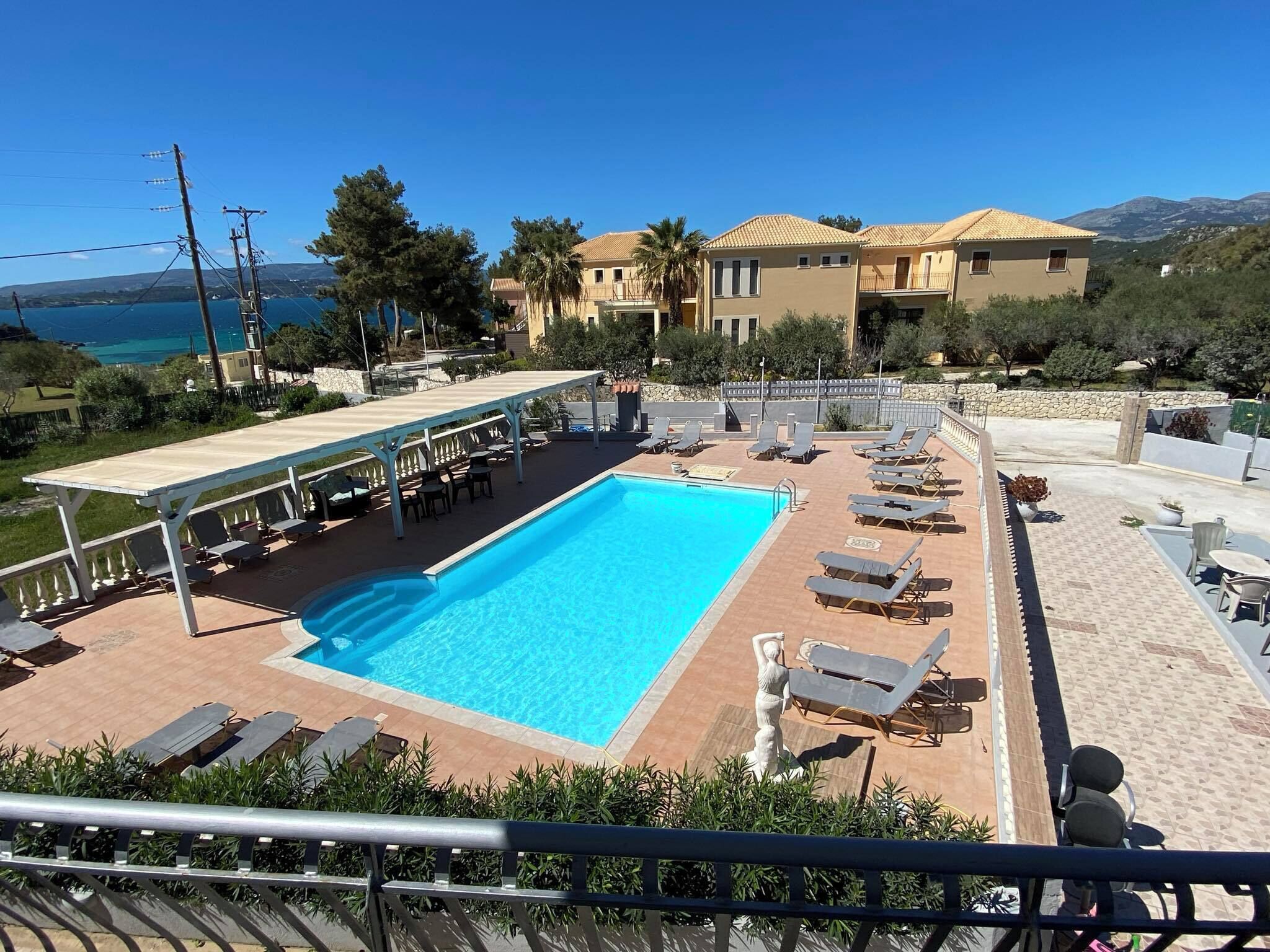 Una piscina con sillones de descanso y una zona cubierta, rodeada de edificios y vista al mar.