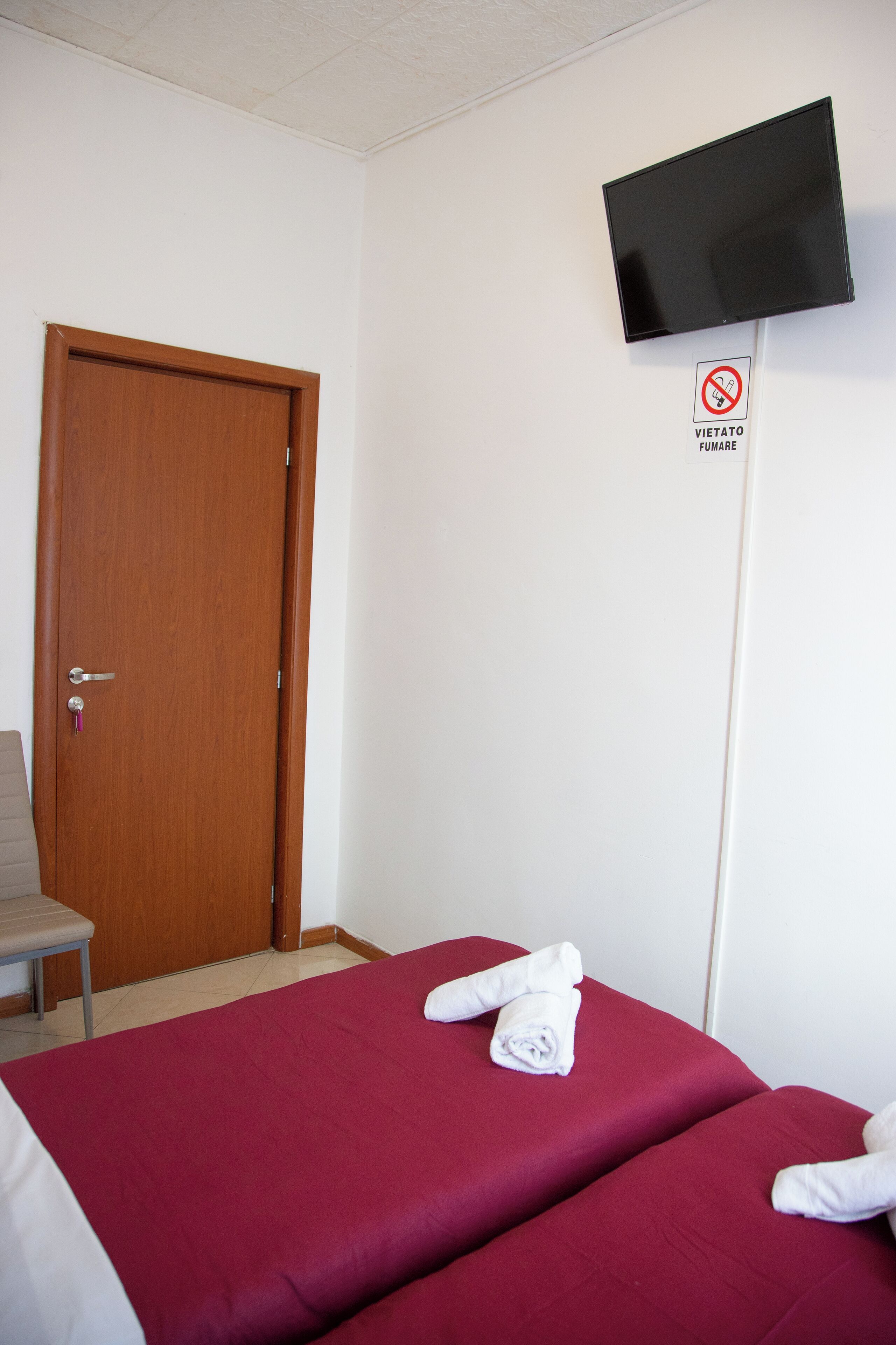 A hotel room with a wooden door, a television mounted on the wall, and a bed with red bedding and rolled towels.