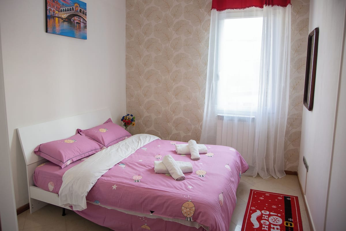 A neatly made bed with pink bedding and white pillows, a window with sheer curtains, and a red and white decorative rug on the floor.