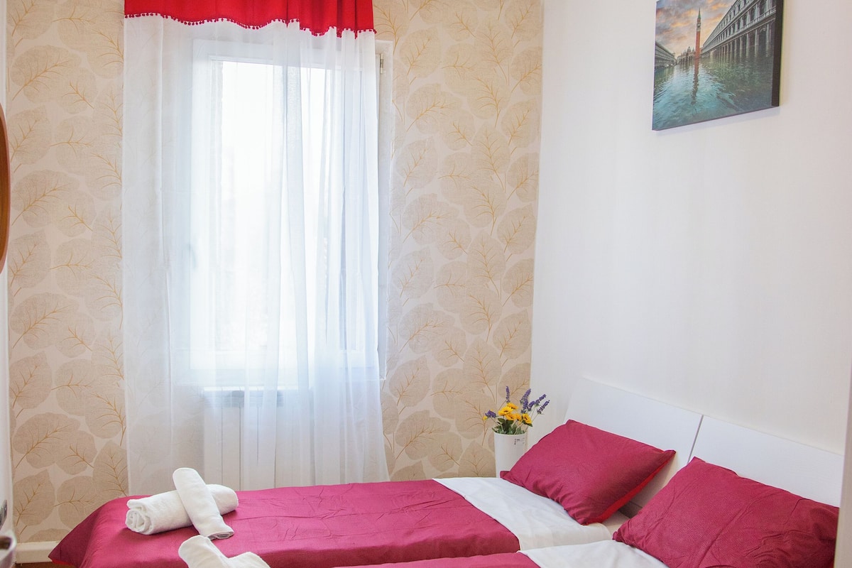 A hotel room with two beds, each with red bedding and white pillows, a window with sheer curtains, a painting on the wall, and a vase with flowers on the nightstand.