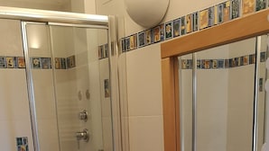 Un baño con ducha envuelta en vidrio, tocador de madera con un lavamanos grande y un espejo sobre el tocador.