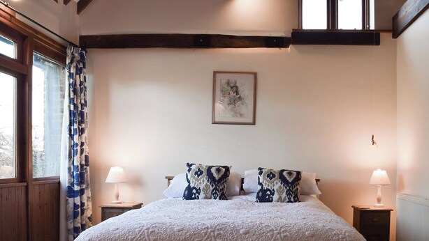 A bedroom with a large bed, two bedside lamps, a window with curtains, and a framed picture on the wall. - Byre Cottage - Meadowbrook Farm (Moreton, near Thame)