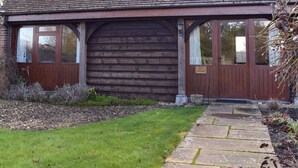 A wooden cottage with a large door and windows, a stone pathway leading to it, and a sign indicating it is a 'BYRE COTTAGE'. - Byre Cottage - Meadowbrook Farm (Moreton, near Thame)