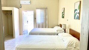 Hotellihuone, jossa on kaksi sänkyä, ovi, joka johtaa käytävään, ikkuna, jossa on verhot, ja seinälle asennettu ilmastointilaite.