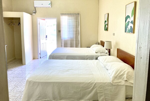 A hotel room with two beds, a door leading to a hallway, a window with curtains, and a wall-mounted air conditioner. - THE IRIE INN APT #2 (Montego Bay)