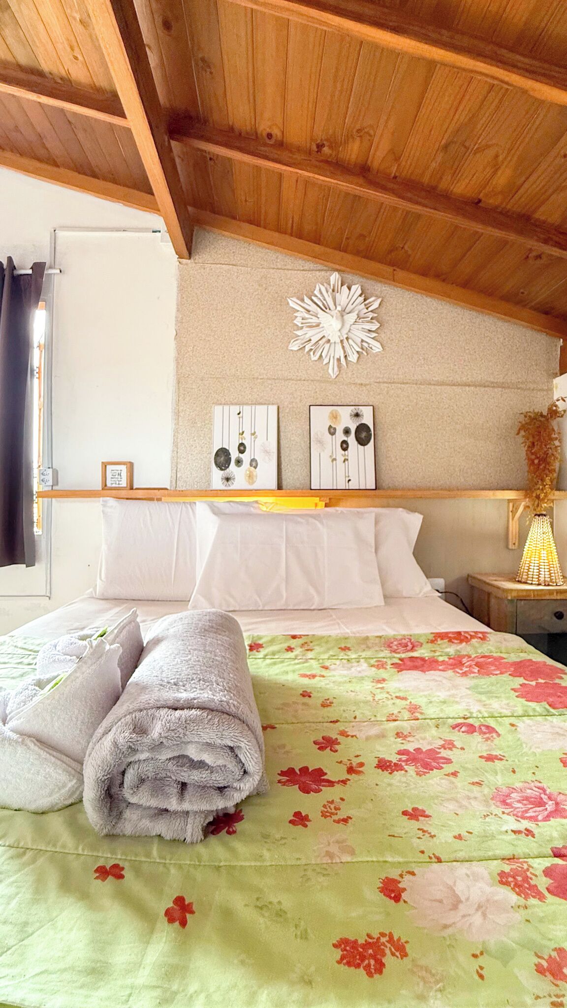 A bedroom with a wooden ceiling, a bed with a floral bedspread, white pillows, and a decorative wall piece.