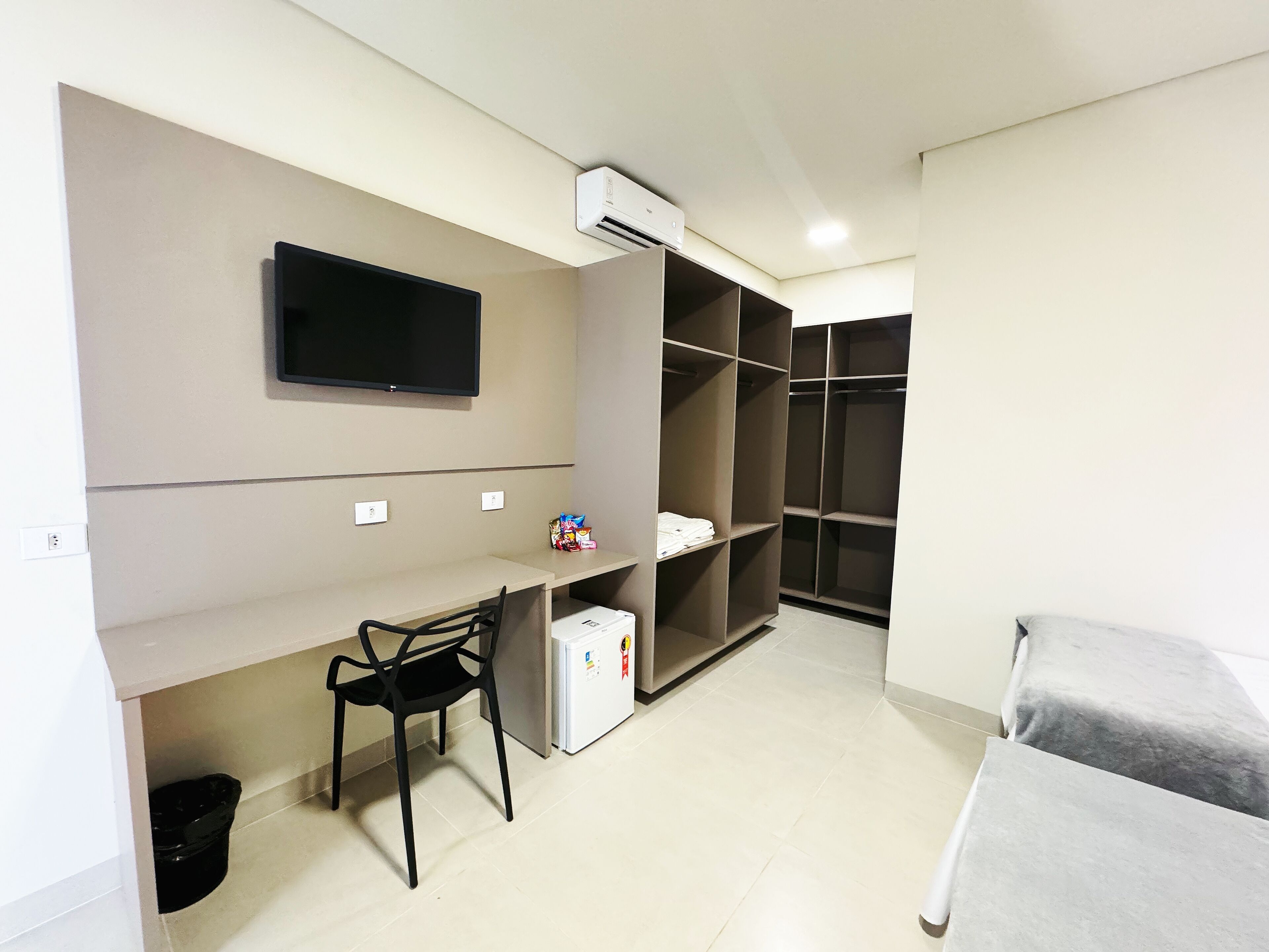 Quarto com cama, escrivaninha com cadeira, televisão fixada na parede e guarda-roupa.