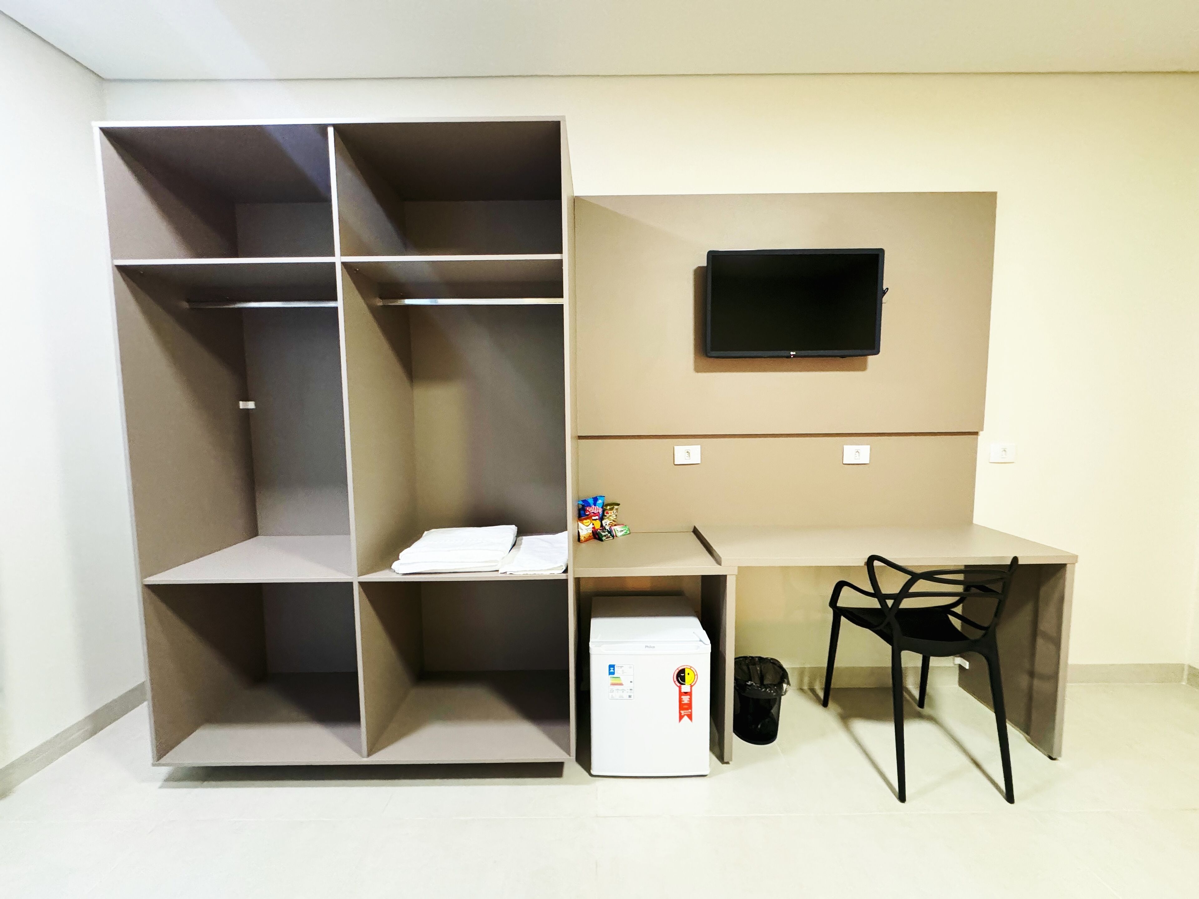 Quarto compacto com roupeiro embutido, escrivaninha com cadeira, pequeno frigorífico e televisão montada na parede.