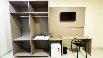 Quarto compacto com roupeiro embutido, escrivaninha com cadeira, pequeno frigorífico e televisão montada na parede.