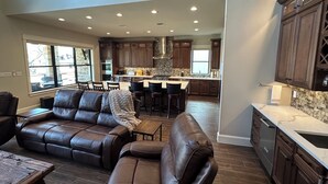 A spacious living room with a leather sofa and a dining area with a kitchen in the background. - New Lake Paradise! 4 bdrm 3.5 ba house w/ lift, deck, grill, sauna, smart, media (Tool)