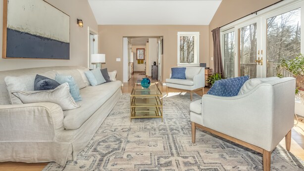 A spacious living room with a beige sofa, a white armchair, and a glass coffee table. There is a large abstract painting on the wall, a ceiling fan, and a view of the outdoors through large windows.