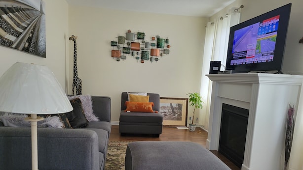 A living room with a grey sofa, a grey ottoman, a small side table with a lamp, a fireplace, and a television displaying news. - Jack and Jill private suite, safe and relaxing 2 bedroom apartment @ RoseGlen  (Niagara-on-the-Lake)