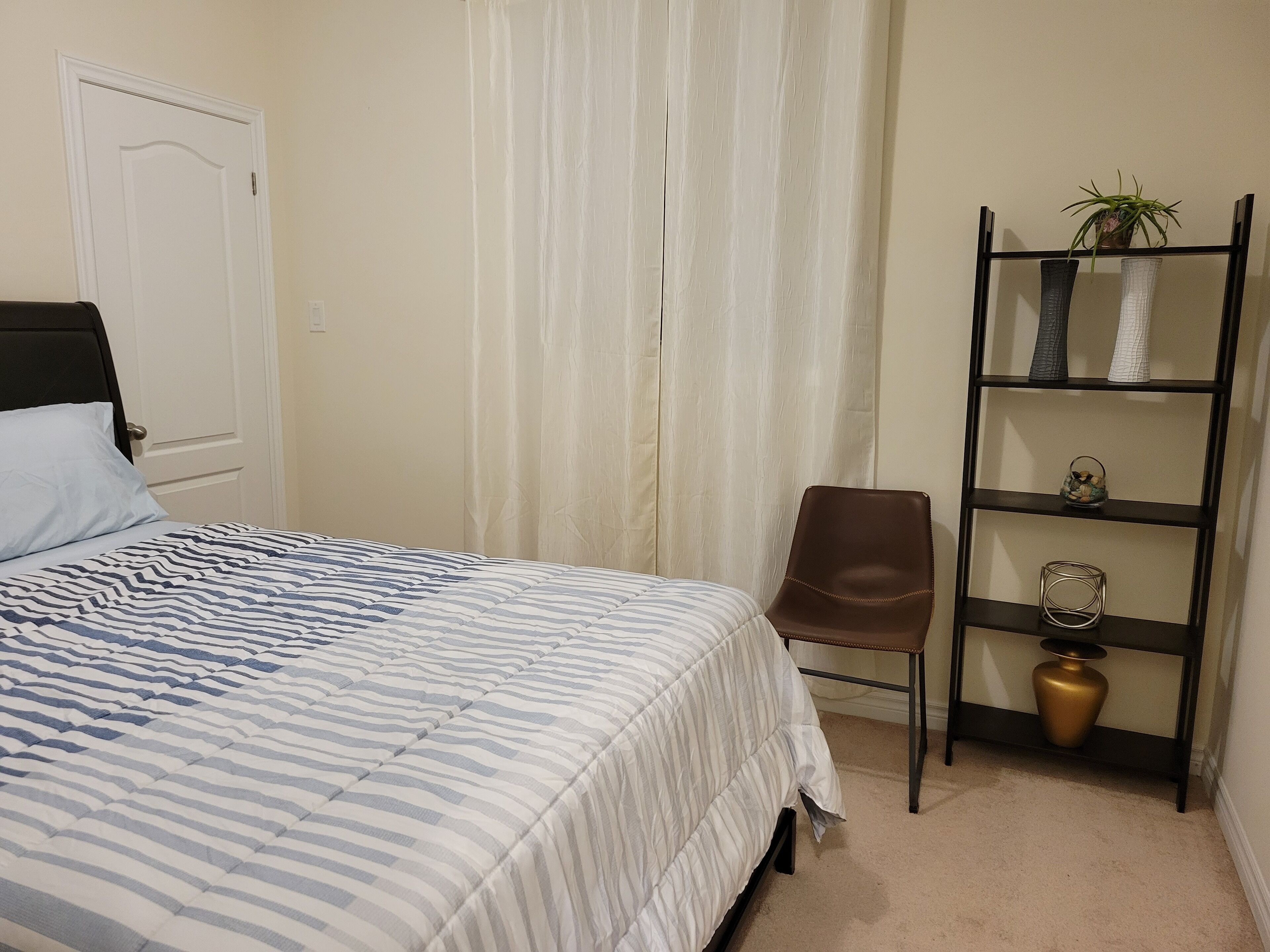 Un dormitorio con cama, una silla, un estante con objetos decorativos y una puerta.