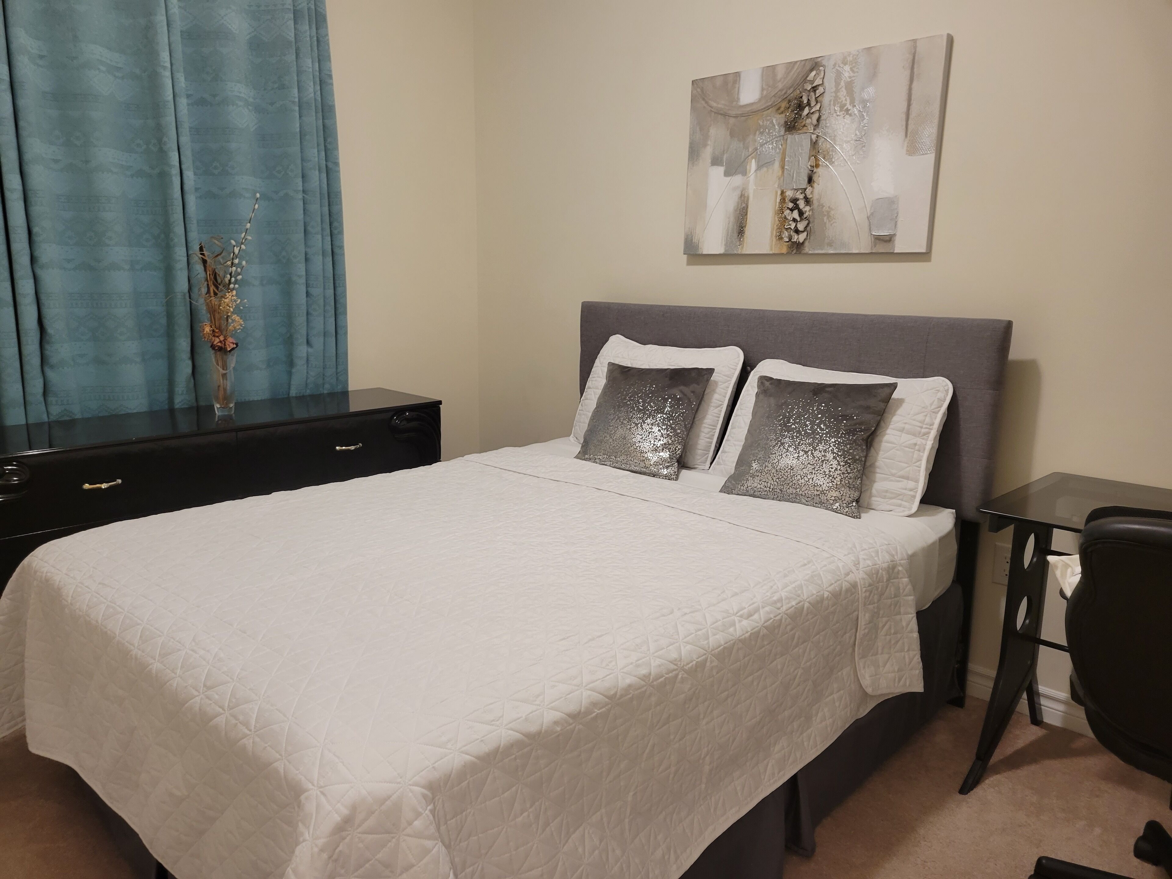 A neatly made bed with a white quilted bedspread, two pillows, a small bedside table, and a wall-mounted abstract painting.