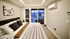 A bedroom with a large bed, a view of the outdoors through a sliding door, and an air conditioning unit on the wall.