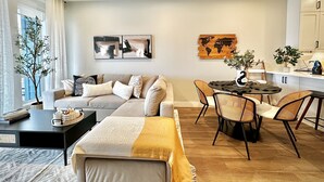 A modern living room with a sofa, coffee table, and dining area. There are two paintings on the wall, a potted plant, and a view of the city through large windows.