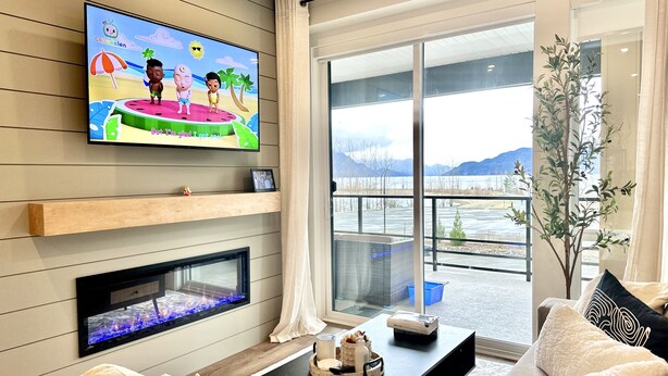 A modern living room with a flat-screen TV, fireplace, comfortable seating, and a large window offering a view of a lake and mountains.