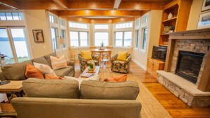 A cozy living room with a fireplace, comfortable seating, and large windows offering a view of the outdoors.