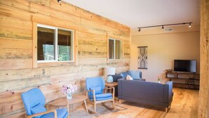 L’intérieur d’un chalet accueillant, avec des murs en bois, un canapé, deux fauteuils bleus, une petite table sur laquelle est posé un vase de fleurs et un téléviseur.