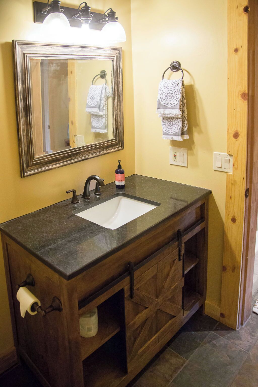 A bathroom with a wooden vanity, a white sink, a mirror with lights, and a towel rack.