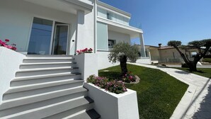 Une maison moderne à deux étages, avec une façade blanche, un escalier menant à l’entrée et un jardin bien entretenu agrémenté de fleurs et d’un petit arbre.