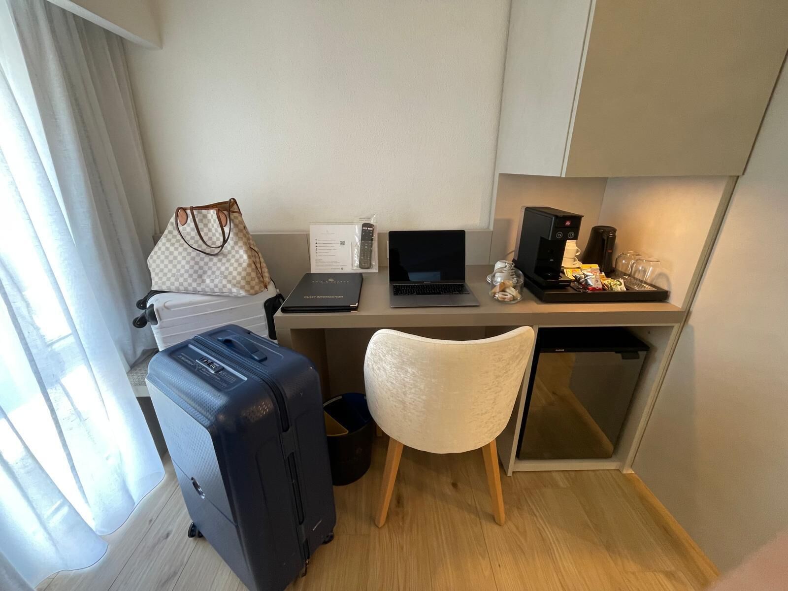 Una habitación de hotel compacta con escritorio, laptop, cafetera y maleta.