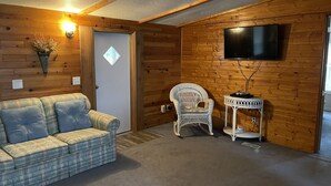 Une pièce chaleureuse avec un canapé à carreaux, un fauteuil en osier, une petite table avec une lampe et un téléviseur à écran plat fixé au mur.