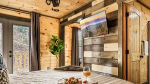 Une pièce chaleureuse, aménagée dans une cabane en bois, avec un lit, un plateau contenant des gaufres et un verre de vin, un tableau accroché au mur et une vue sur l’extérieur.