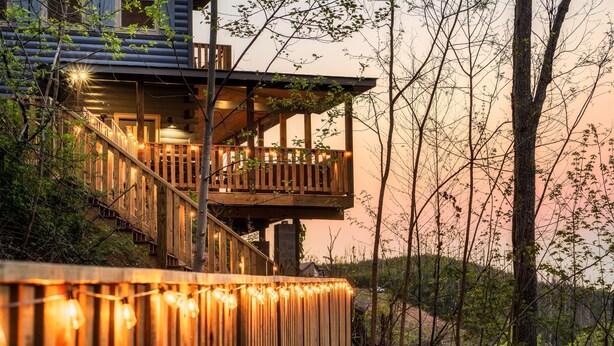 Une structure en bois à plusieurs niveaux, dotée de balcons, de guirlandes lumineuses et entourée d’un cadre naturel.