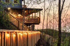 Une structure en bois à plusieurs niveaux, dotée de balcons, de guirlandes lumineuses et entourée d’un cadre naturel.