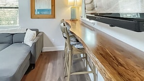 A modern living room with a wooden ceiling, a grey sofa, a dining table with chairs, a large window, and a TV.