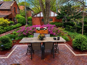 A patio with a wooden table and chairs, surrounded by lush greenery and flowering plants.