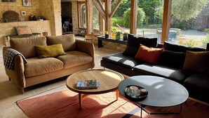 A cozy living room with a stone wall, wooden beams, and large windows overlooking a garden. - Maison Nina: un Ancien Corps de Ferme Rénové Avec Piscine, Entre Dordogne et Lot (CAMPAGNAC LES QUERCY)