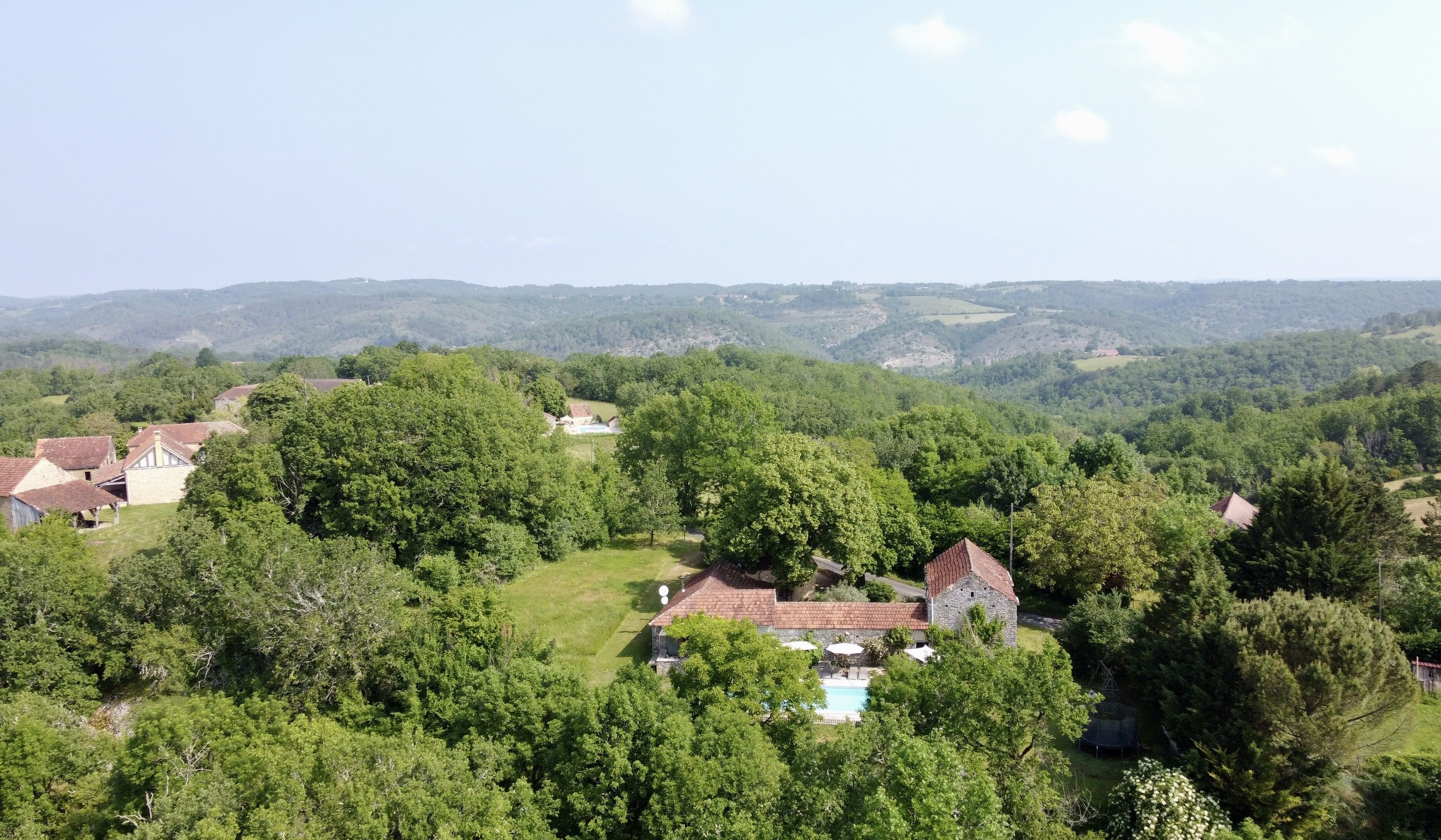 A rural landscape with a house surrounded by dense greenery and hills in the background.
