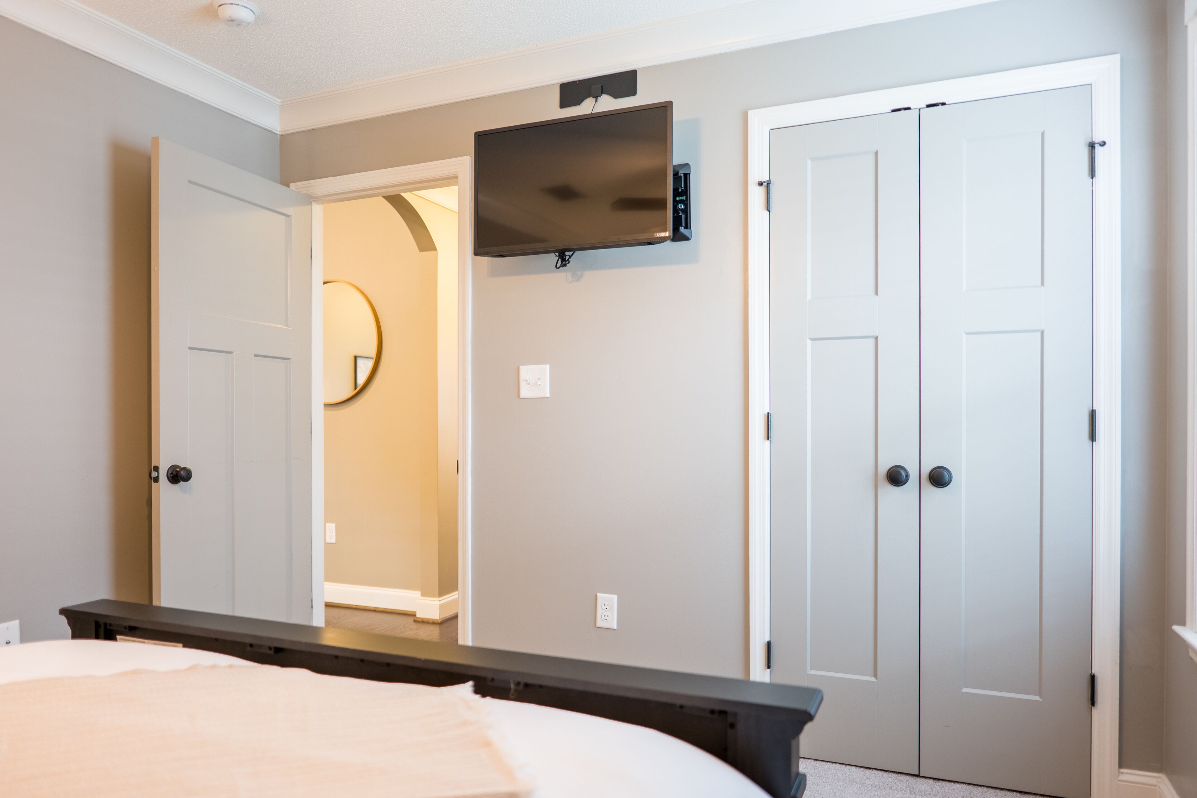 A bedroom with a bed, a flat-screen TV mounted on the wall, a wardrobe, and a door leading to another room.