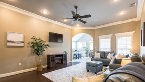 A spacious living room with a ceiling fan, a flat-screen TV mounted on the wall, a large potted plant, and a grey sofa with multiple cushions. - Peaceful Retreat on Newbury with Pool, Soaking Tub, King Bed (Valdosta)