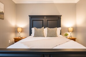 Ein ordentlich bezogenes Bett mit zwei Kissen, an beiden Seiten mit Nachttischen und Lampen, in einem Zimmer mit dunklem Kopfteil und hellen Wänden.