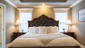 A neatly made bed with a tufted headboard, two bedside tables with lamps, and a window with blinds. - Peaceful Retreat on Newbury with Pool, Soaking Tub, King Bed (Valdosta)
