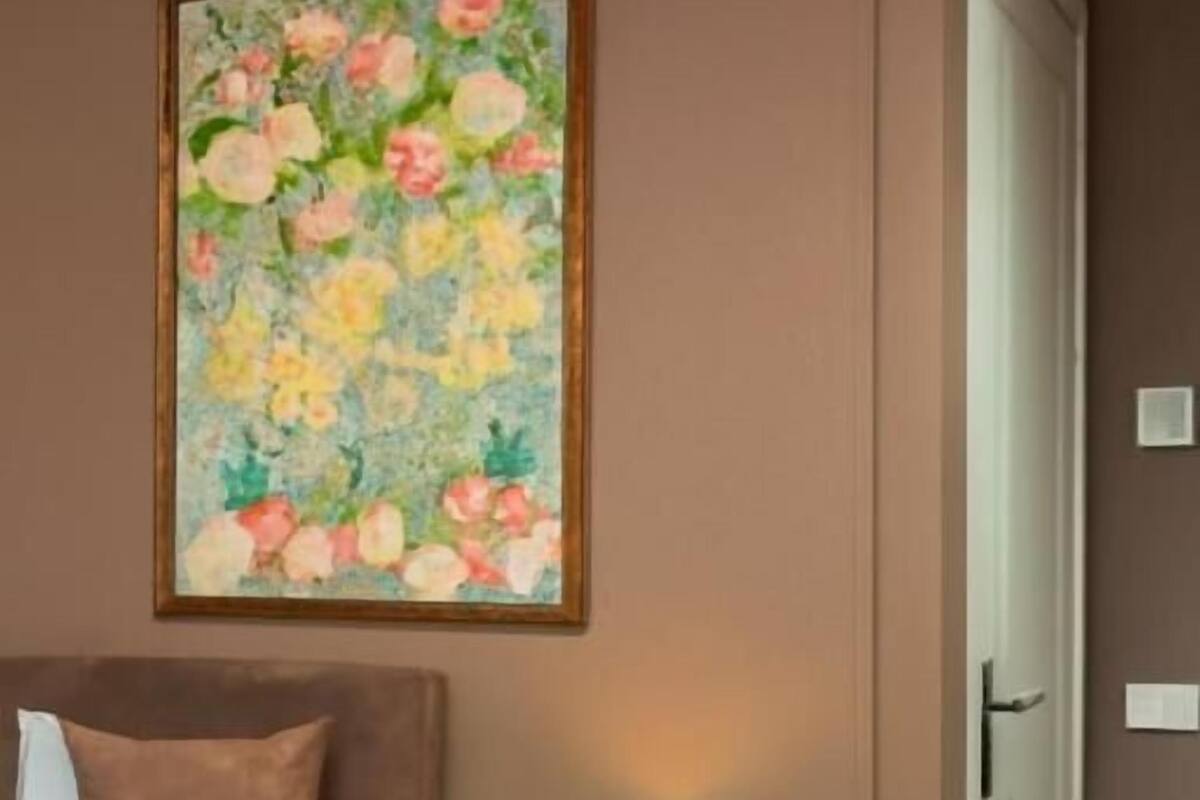 A hotel room with a bed, a blue lamp on a nightstand, a painting of flowers on the wall, and a door.