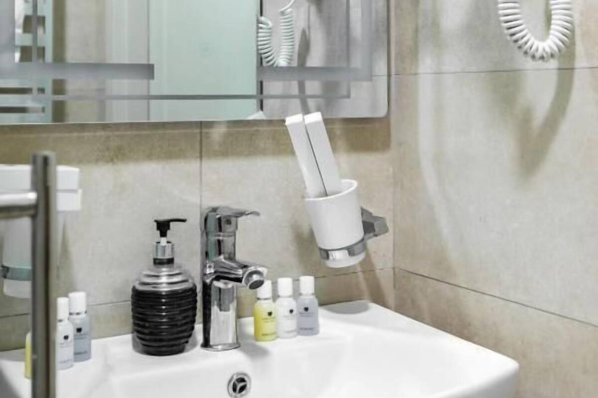 A bathroom with a white sink, a mirror, a hairdryer mounted on the wall, and toiletries on the sink.