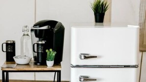 A kitchenette with a white refrigerator, a coffee maker, a glass carafe, a bowl, and a small potted plant on a wooden table.