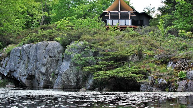 Une maison avec un toit triangulaire, entourée d’une forêt dense et d’un terrain rocheux.